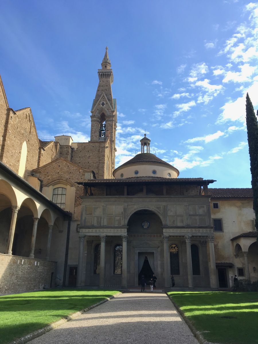 Courtyard-like area at Santa Croce, featuring a gravel pathway and green grass. Surrounding the open space are covered walkways made of grey and brown stone. In the background, a steeple rises into the blue sky.