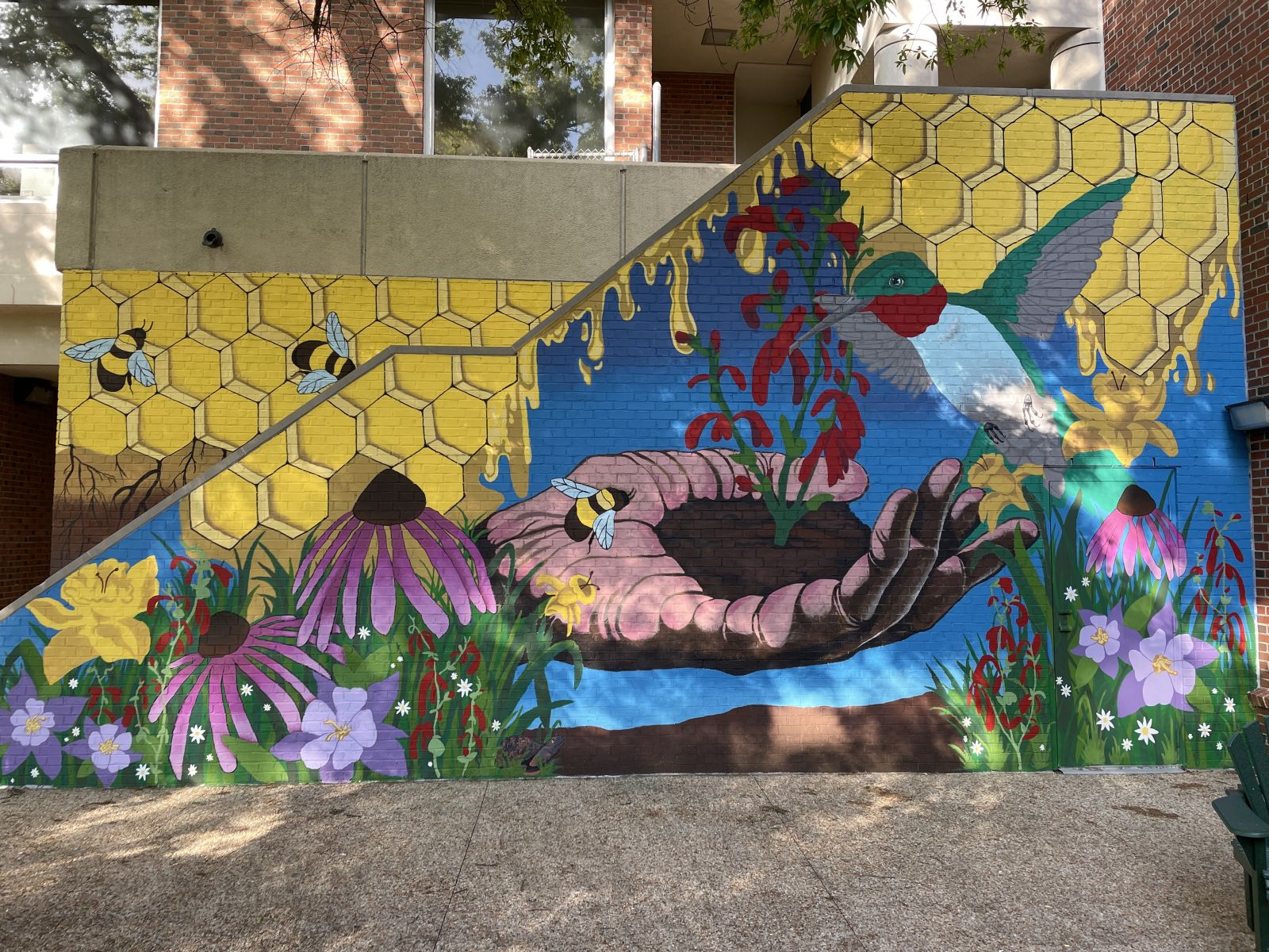 view of a painted mural with a hand holding a red cardinal flower plant with soil, surrounded by native plants and pollinators