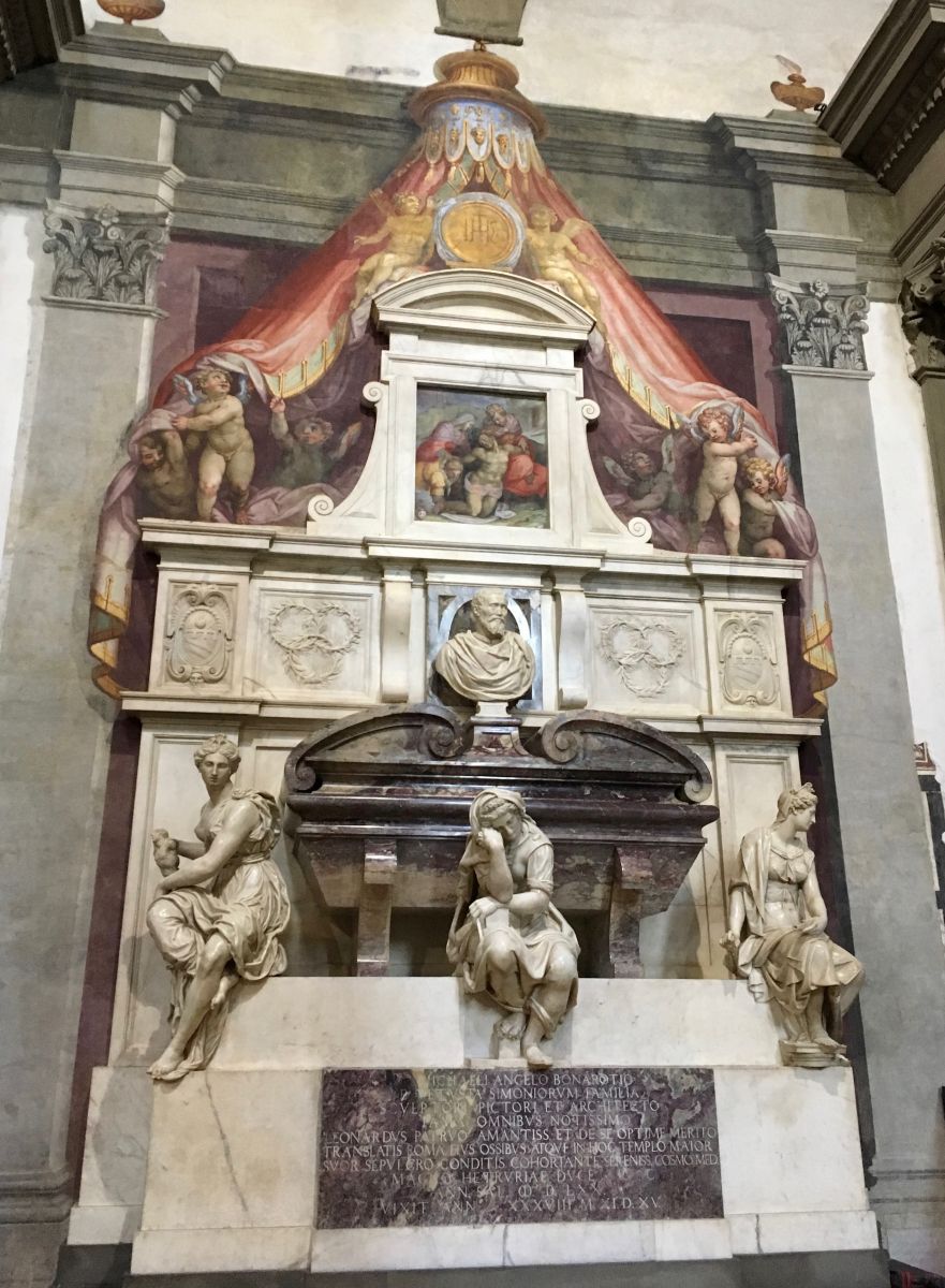 Ornate tomb that appears to be made of marble. Three figures sit towards the bottom, while a bust of a man (Michelangelo?) rests above them. Pieta imagery and cherubs are painted above the marble tomb's mantle. Text written in Latin is inscribed towards the bottom of the tomb, near the floor.