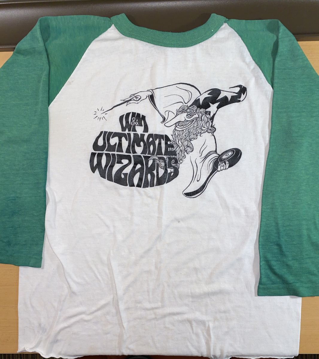 White Ultimate Frisbee jersey shirt with light green sleeves. In the center of the shirt is a black-and-white graphic of a wizard holding a wand in one hand and a frisbee in another. The shirt reads, "W&M / Ultimate / Wizards."