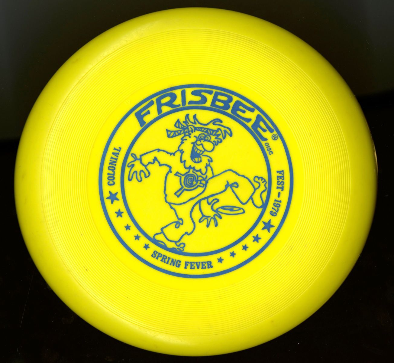 Yellow frisbee with blue text and a blue line illustration of a "hippie" caricature. The disc reads, "Colonial / Fest - 1979 / Spring Fever." 