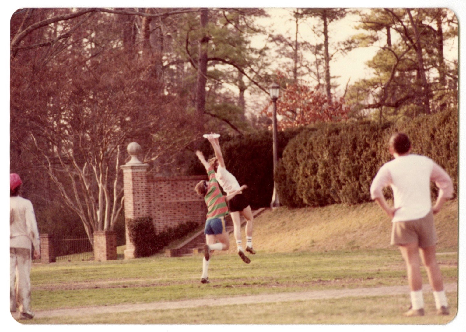 Photograph from the 1980s featuring two Ultimate Frisbee players reaching for a flying disc on the Sunken Garden. Another two players stand with their backs to the camera, watching the contest take place.