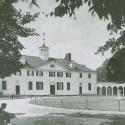 A black and white photo of the Mansion at Mount Vernon is taken from some trees to the left of the house. A man kneels, and a young girl stands beside him in the foreground of the picture.